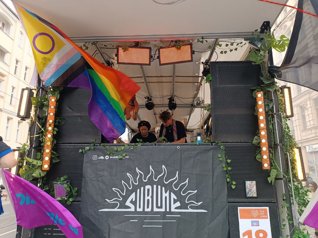 Zwei DJs am Pult auf dem sublime-Truck, eingerahmt von Lautsprechertürmen, Lichterleisten und Efeu. Links wehen eine Regenbogen- und eine Progress-Pride-Flagge, vorne hängt das sublime-Banner mit Sonnenlogo.